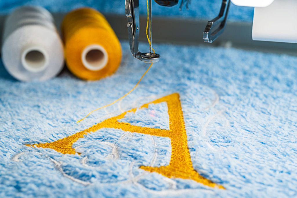 Yellow A alphabet logo design and two color threads on blue towel in hoop of embroidery machine, close up picture