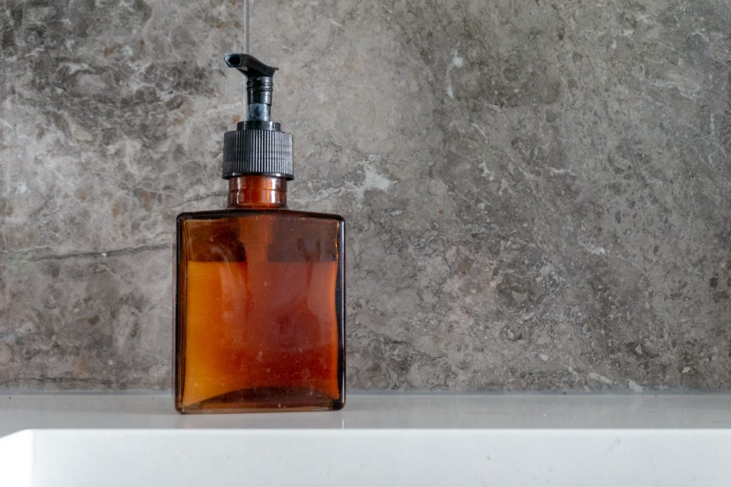 Hand soap dispenser in a brown glass, square bottle, with black pump, and grey marble backsplash near kitchen sink