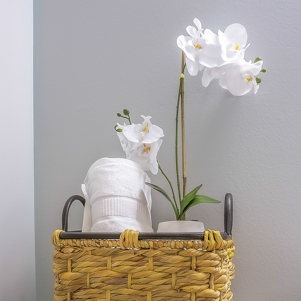 Square Woven basket with tissue roll and white flowers placed on top of the toilet tank. Bathroom sink and mirror can also be seen inside the room.